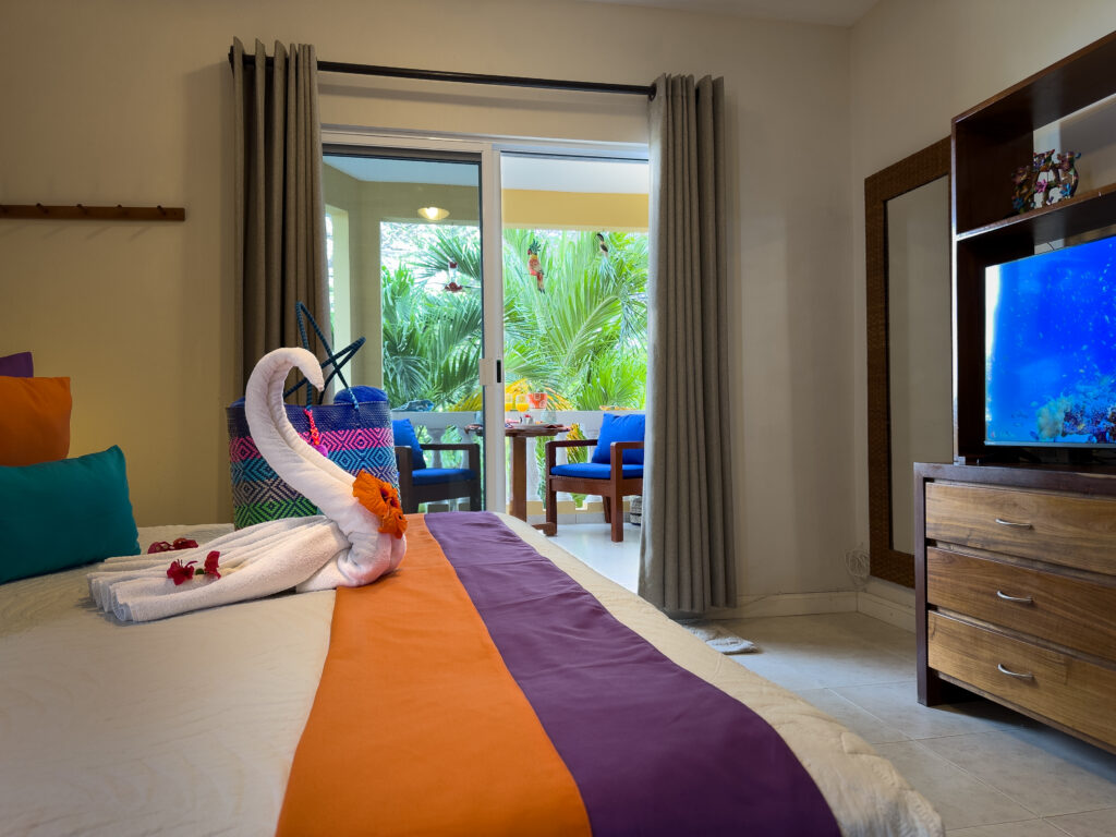 Bed facing a television and sliding doors opening to blue balcony chairs amid palms.
