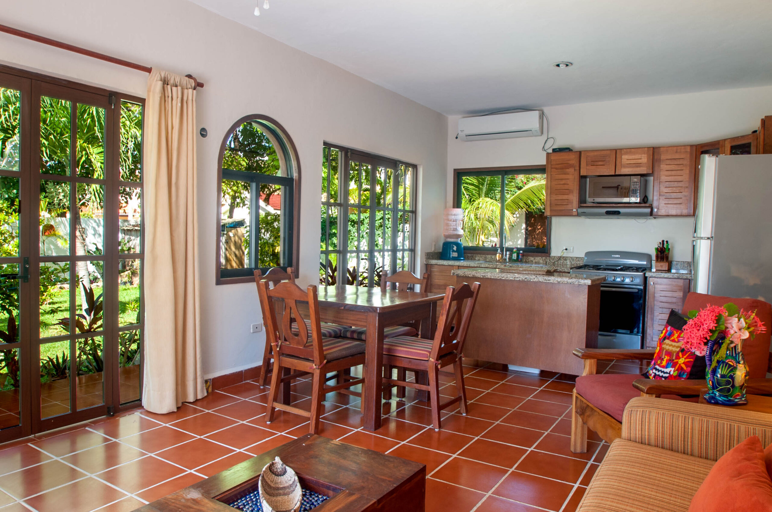 Wooden dining table beside large windows and doors, with the kitchen beyond.