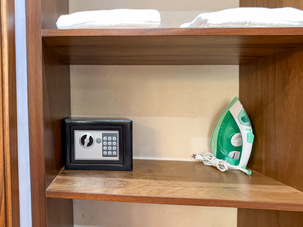Small electronic safe and clothes iron on a wooden shelf beneath folded towels.