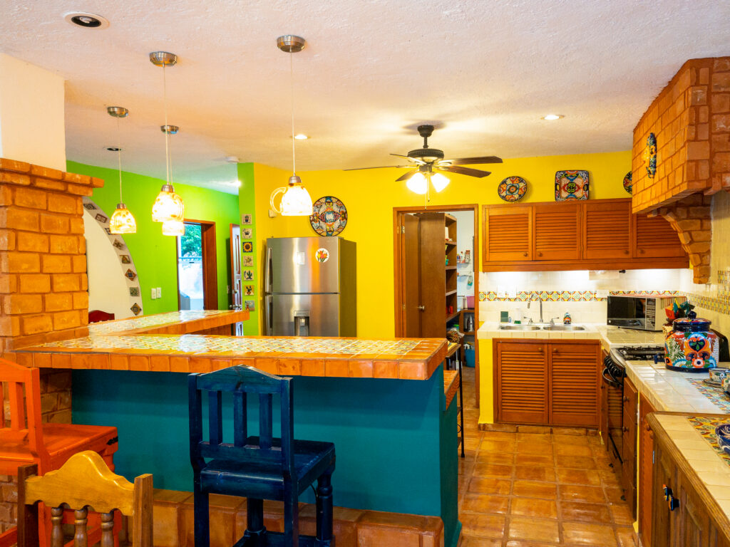 Kitchen with a turquoise island, colorful stools, wooden cabinets, and a stainless steel refrigerator.
