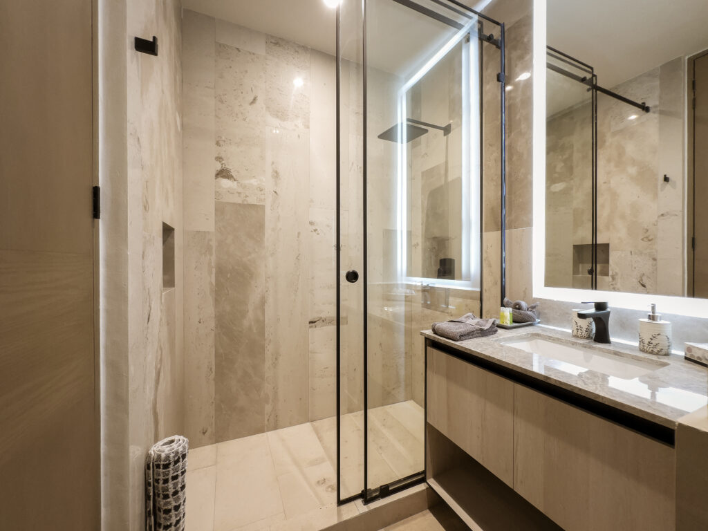 Stone-tiled shower with a glass screen beside a bathroom vanity and illuminated mirror.