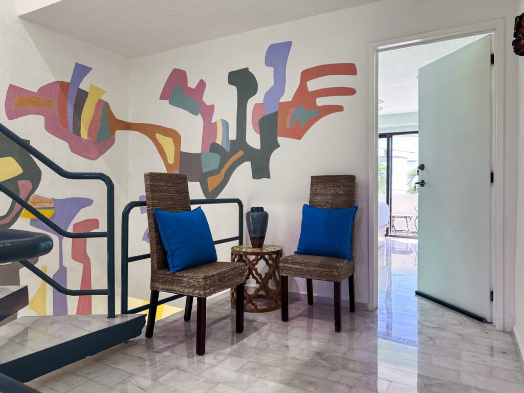 Two chairs with blue cushions beside a small table against a colorful abstract wall mural.
