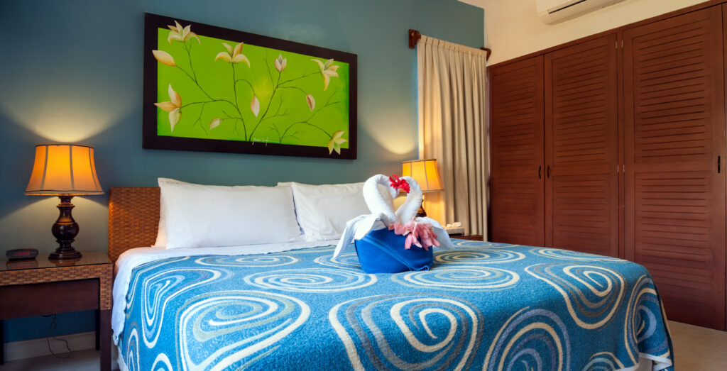 Blue-patterned bedspread and folded towel decoration beneath green floral artwork with lamps on either side.
