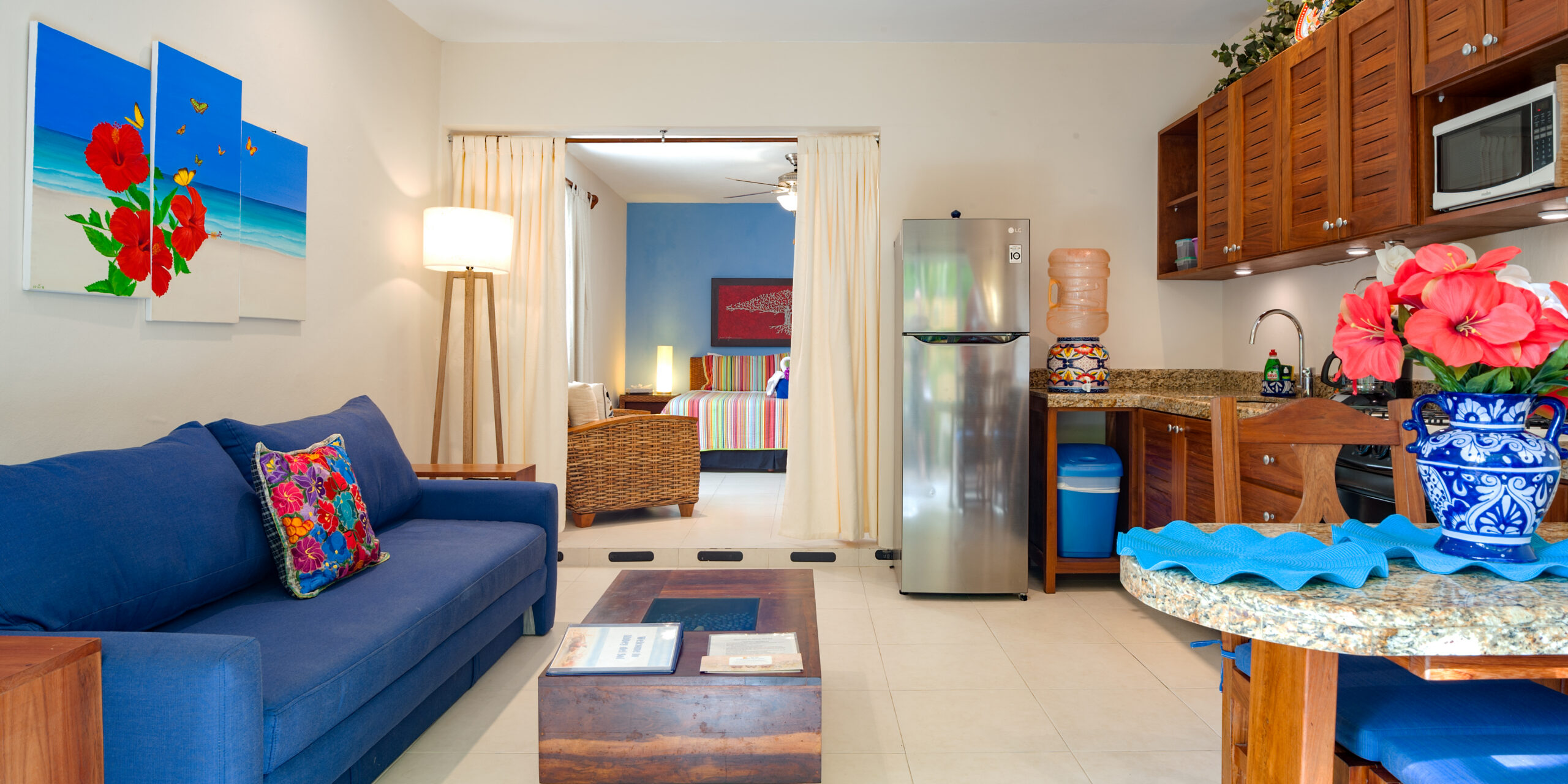 Blue sofa beside a kitchen with wooden cabinets and a stainless steel refrigerator, with the bedroom beyond.