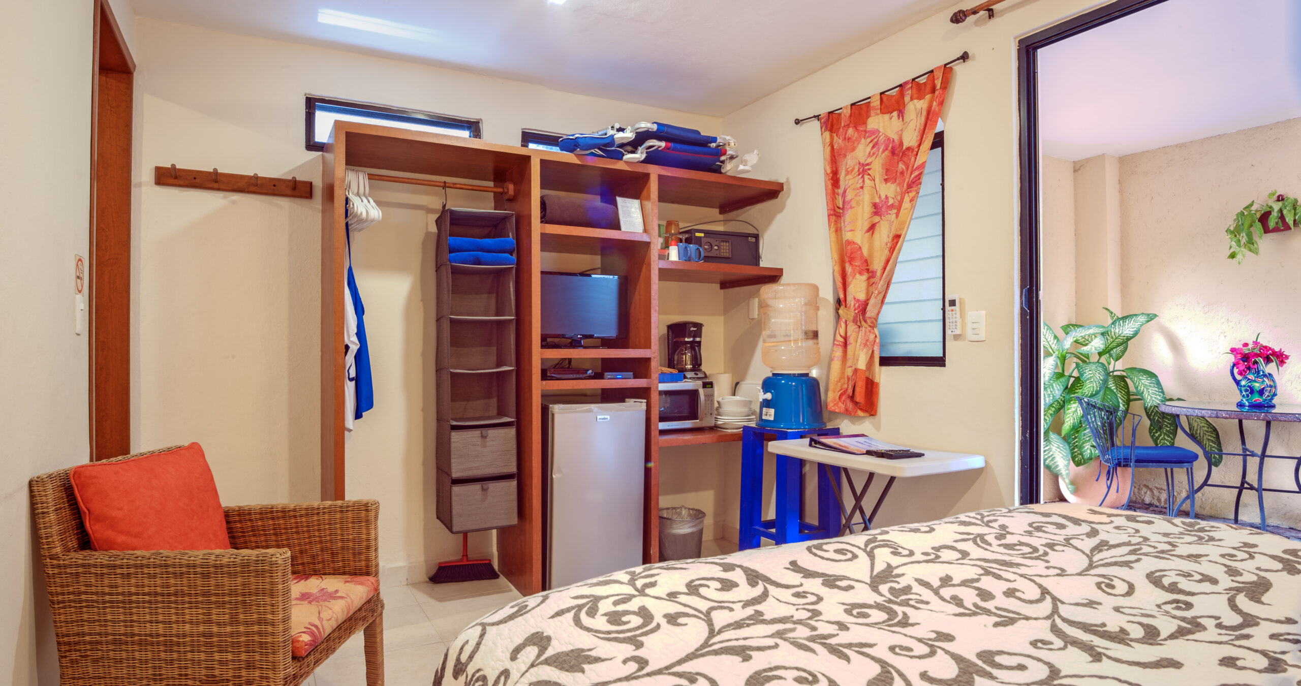 Wooden shelving with a television, small refrigerator, and coffee maker beside the bedroom seating.