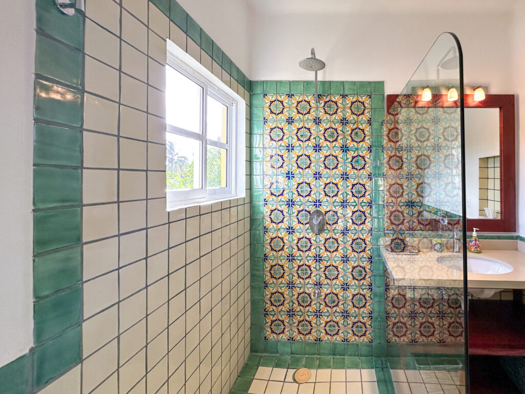 Shower with a colorful patterned tile wall and glass screen beside a window and bathroom vanity.