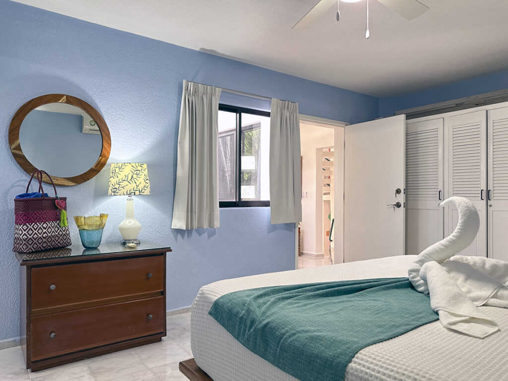 Bed with towel swan facing a dresser, round mirror, and white closet doors.