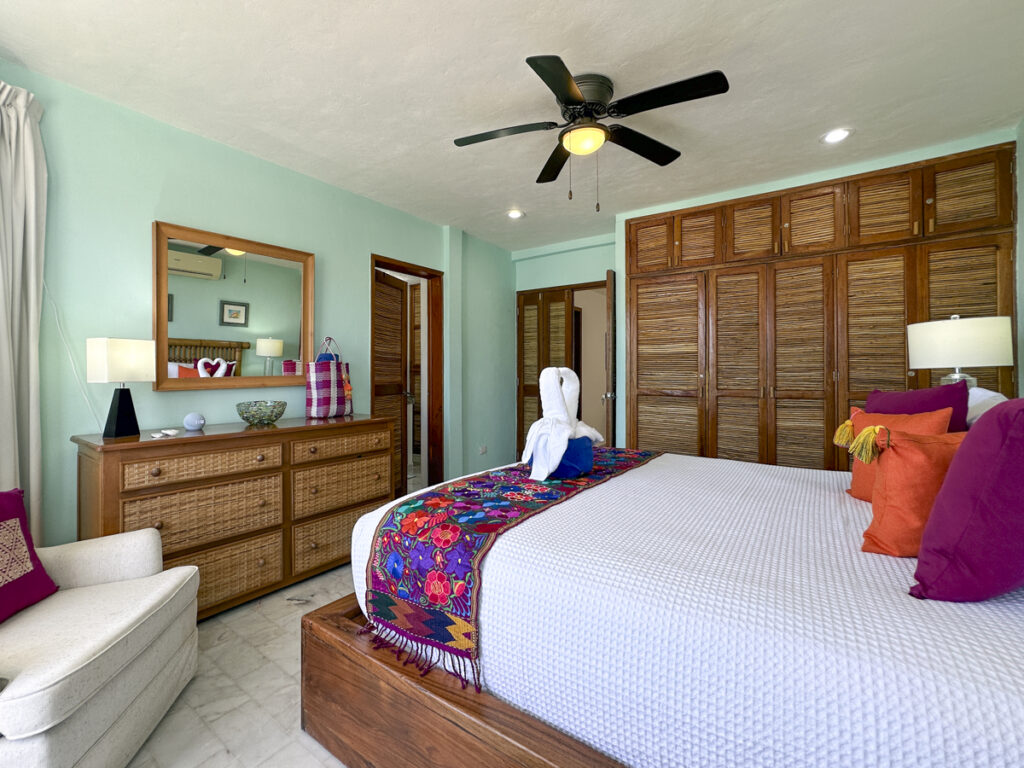 Bed with bright pillows and floral runner beside a dresser and large wooden closet wall.