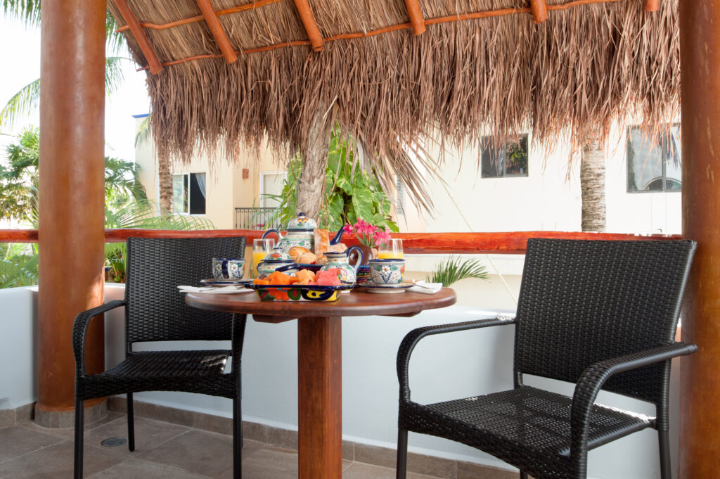Round table set with dishes and food between dark chairs beneath a thatched balcony roof.