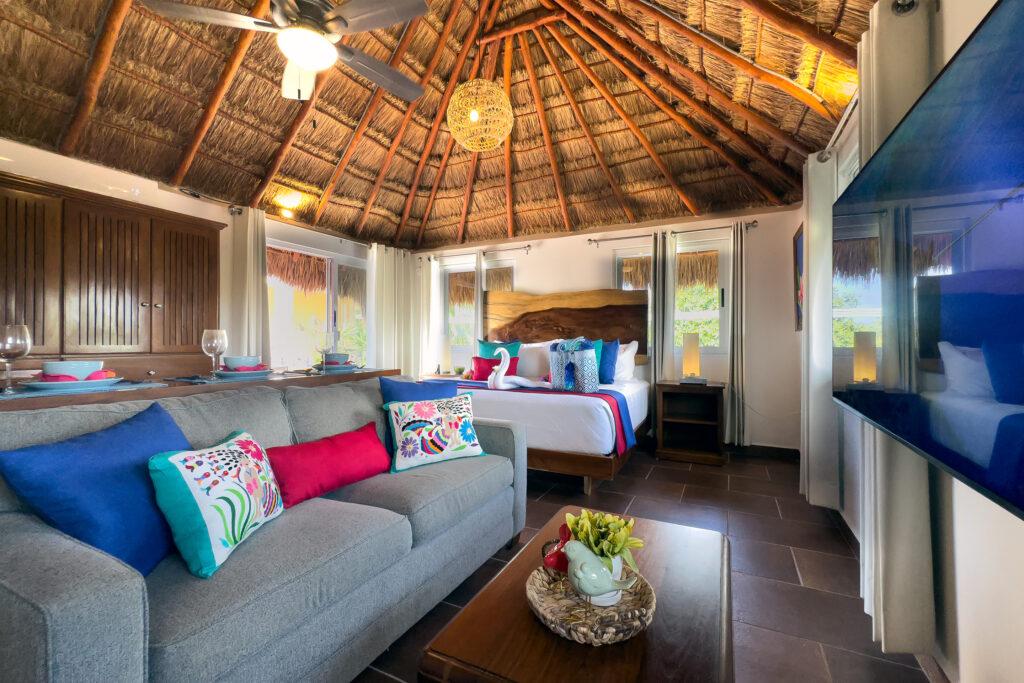 Gray sofa, coffee table, bed, and wall-mounted television beneath a tall thatched roof.