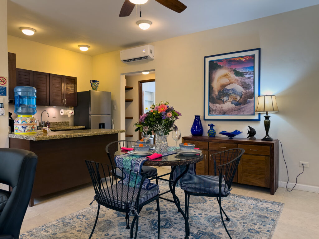 Round dining table with flowers beside a breakfast counter and wooden sideboard beneath framed artwork.