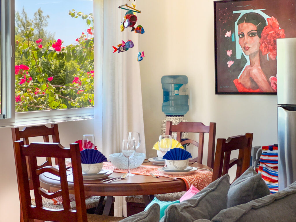 Round dining table beside a window with flowering plants outside and framed portrait artwork.