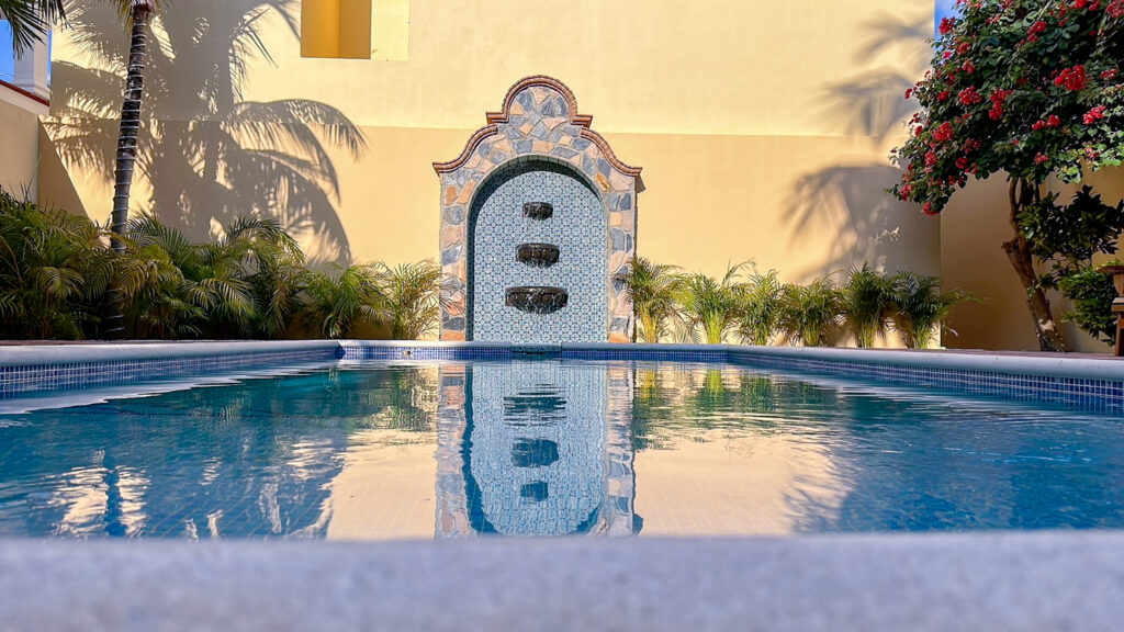 Decorative tiled wall fountain reflected in the blue pool against a yellow garden wall.