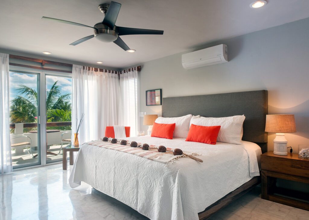 White bed with orange pillows beside glass doors opening onto a balcony with palm views.