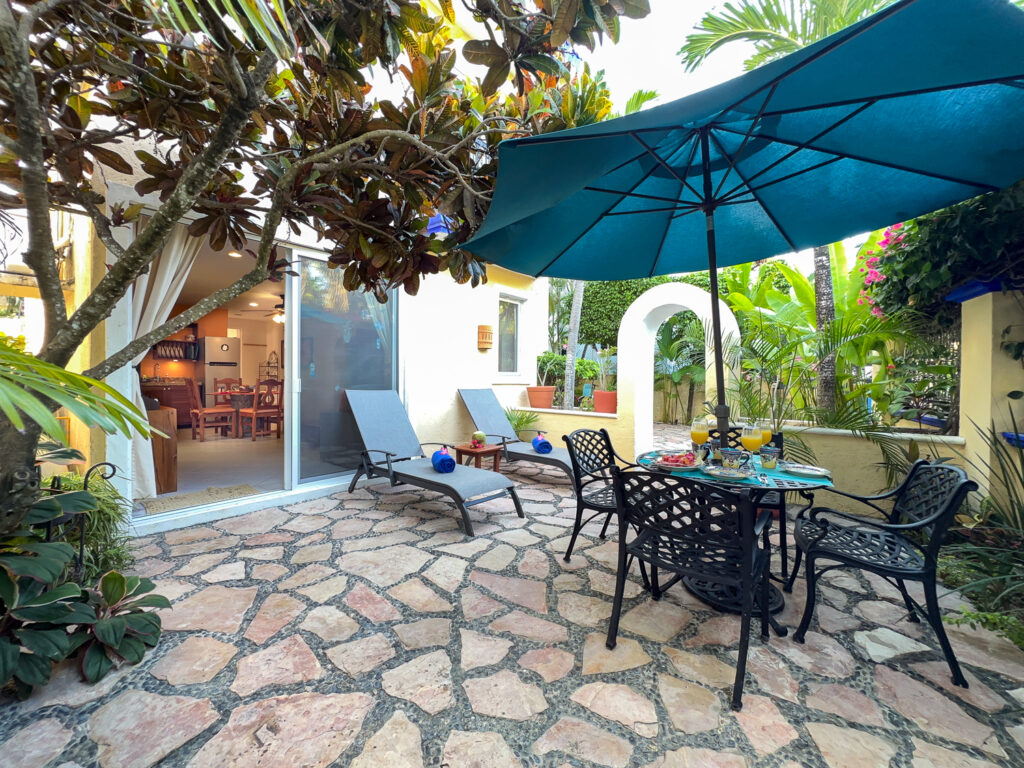 Stone-paved patio with lounge chairs and a dining table beneath a turquoise umbrella among plants.