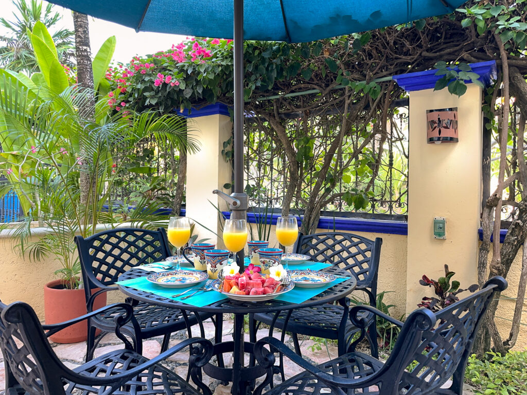 Patio table set with dishes and glasses beneath a turquoise umbrella beside lush greenery.