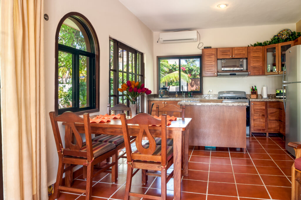Dining table with flowers beside an open kitchen with wooden cabinets and a breakfast counter.