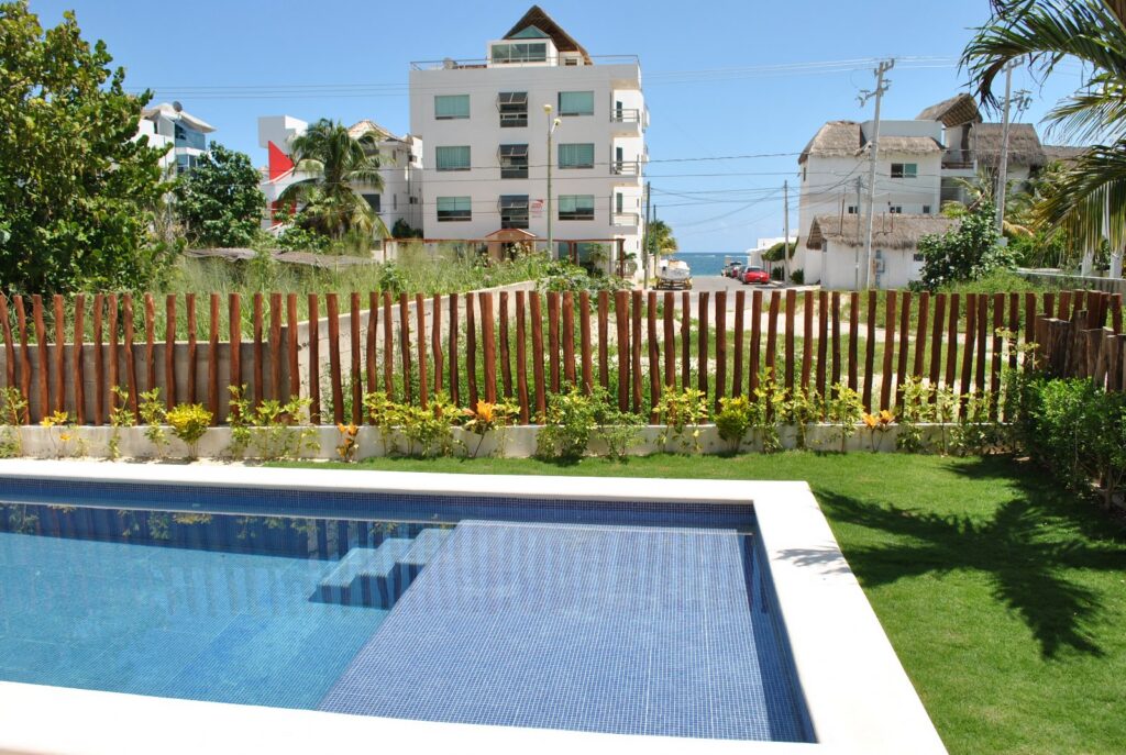 Pool with tiled steps beside a lawn and wooden fence, with nearby buildings beyond.