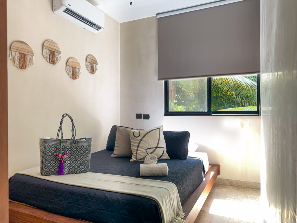Wooden bed with dark bedding, rolled towels, and a woven bag beneath a window with a roller shade.