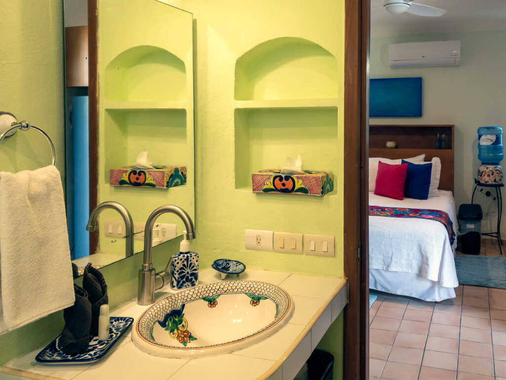 Decorative sink beneath a mirror and green recessed shelves, with the bed visible through the doorway.