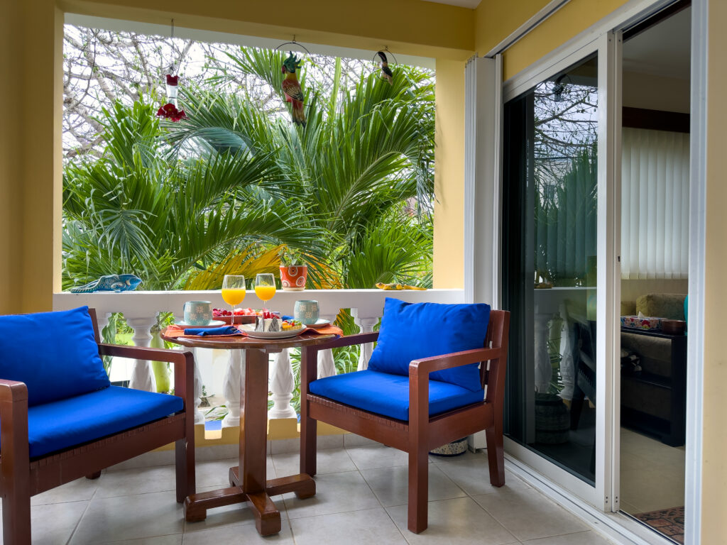 Two blue-cushioned chairs and a table set for two on a covered balcony beside sliding doors.