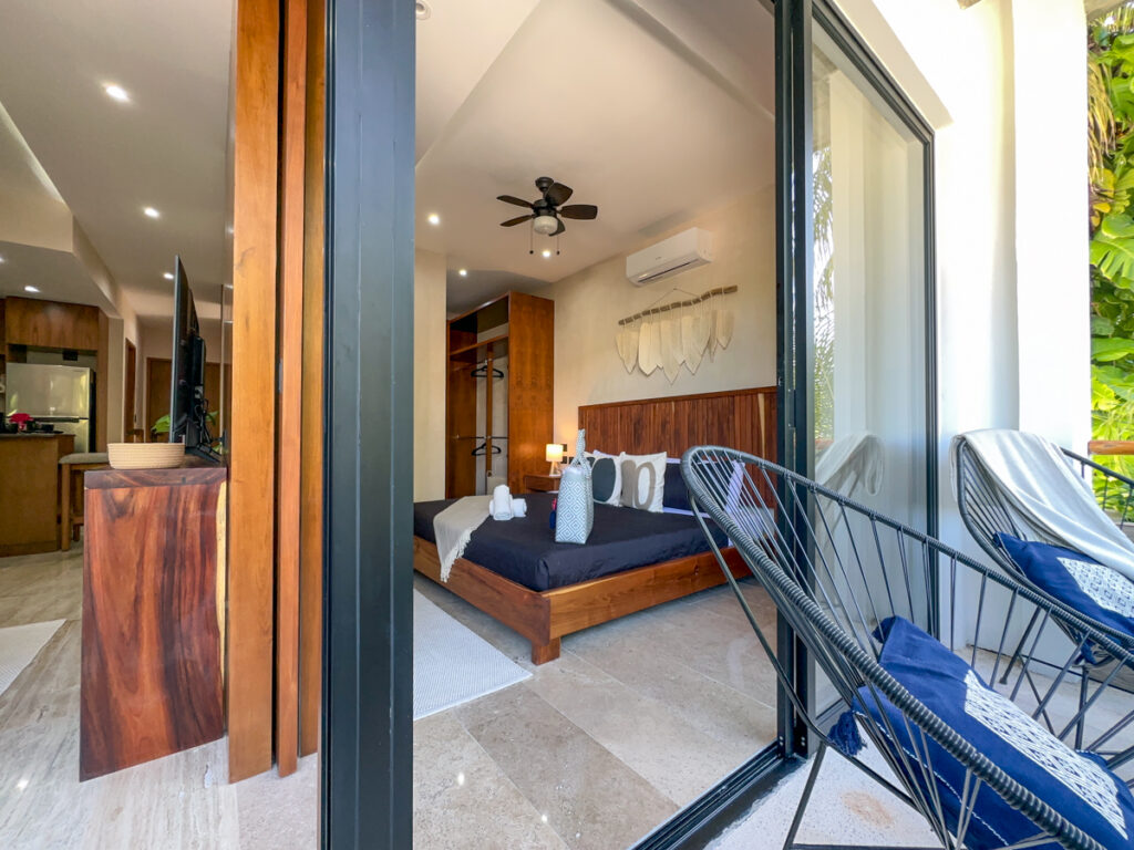 Open glass doors connect a bedroom with dark bedding to a balcony with woven chairs.