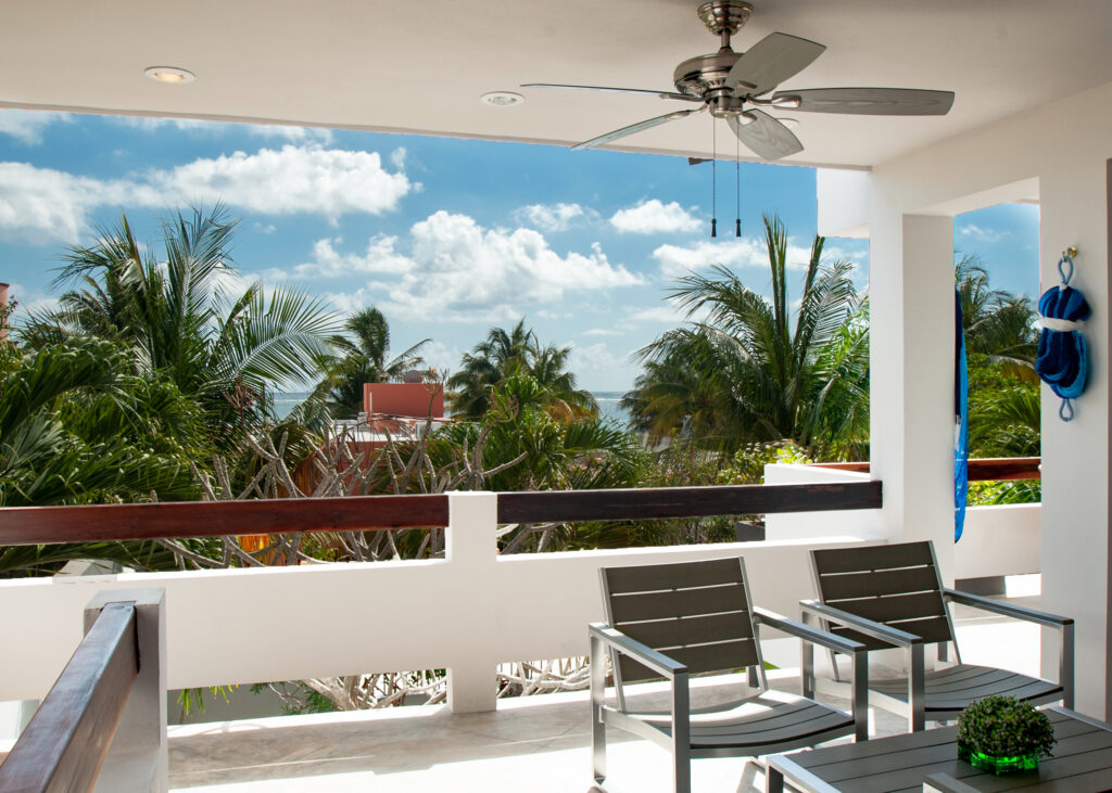 Chairs on a covered balcony overlook palms, rooftops, and the sea beneath scattered clouds.
