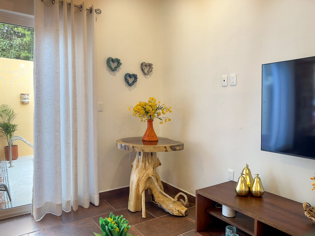 Vase of yellow flowers on a sculptural wood table beside heart wall ornaments and the television.