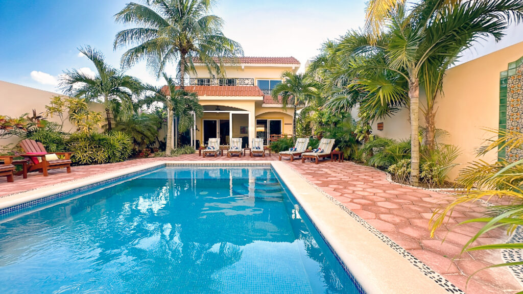 Long blue pool leads toward the yellow house, with palms and lounge chairs along both sides.