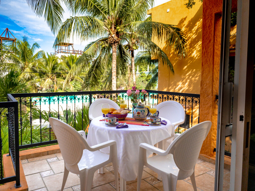 Round table set with dishes beneath a white cloth on a balcony surrounded by palms.