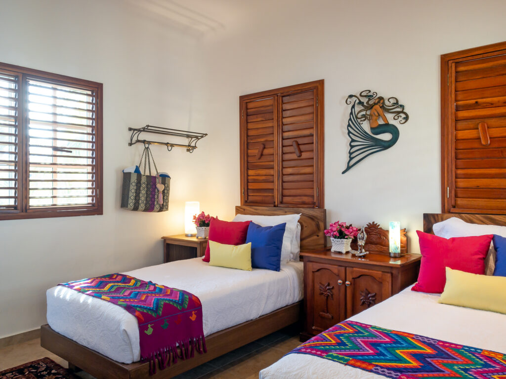 Two single beds with colorful woven runners and bright cushions beneath wall ornaments and wooden shutters.