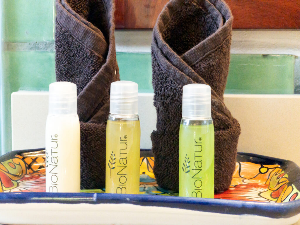 Small toiletry bottles and rolled brown towels arranged on a decorated ceramic tray.