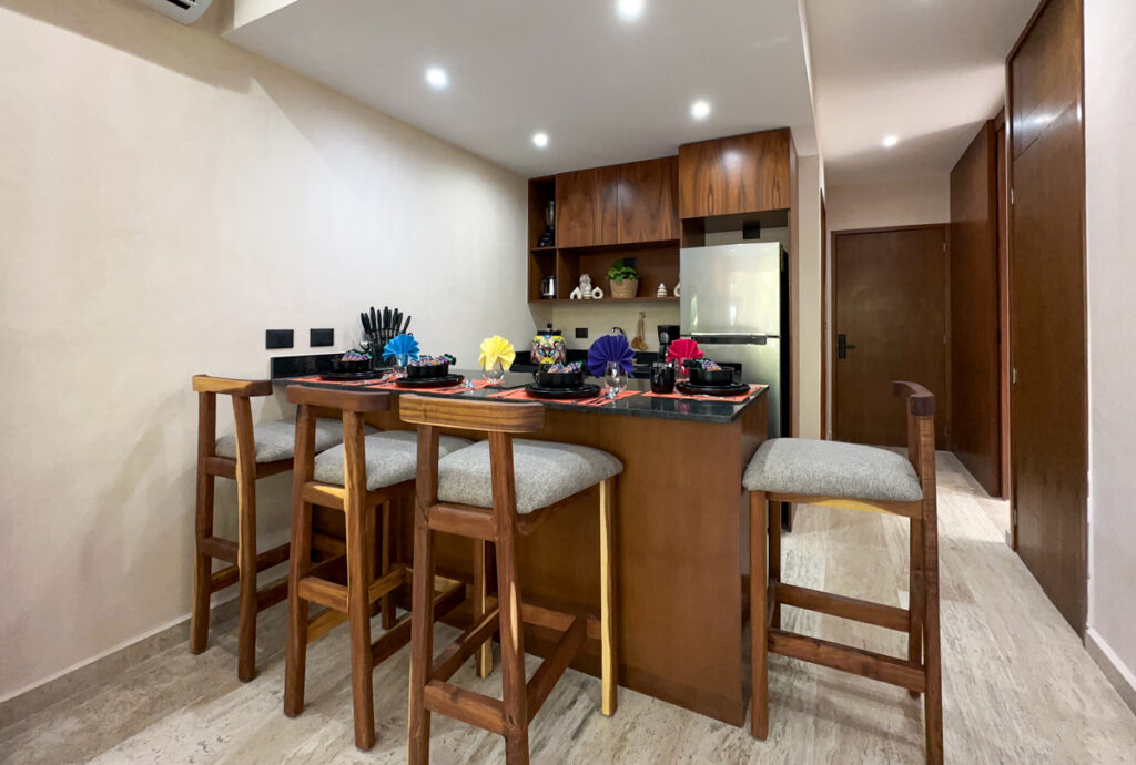Wooden kitchen island set with dishes beside upholstered barstools and a stainless steel refrigerator.