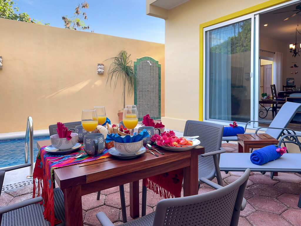 Patio table set with dishes, drinks, and colorful napkins beside lounge chairs and a sliding door.