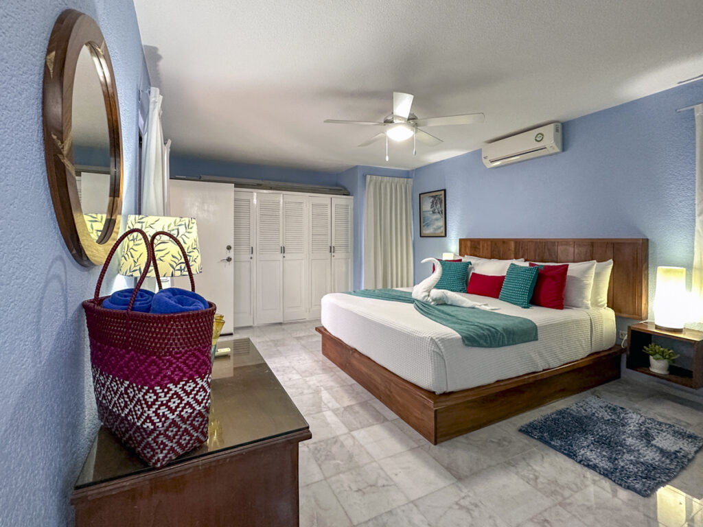 View past a woven bag and round mirror toward a wooden bed with red and green pillows.