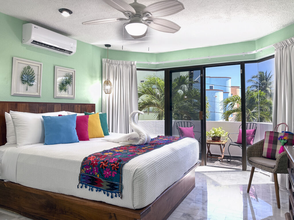 Bed beneath palm-themed pictures beside balcony doors and wicker chairs in a pale green room.