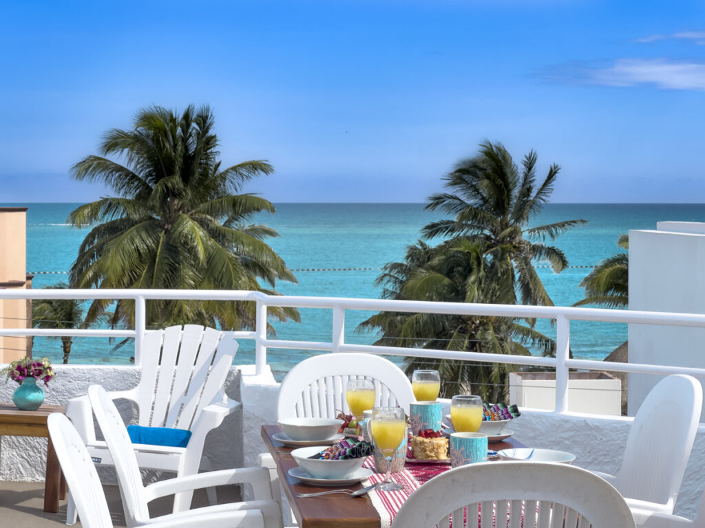 Outdoor table set with dishes and drinks beside white railings overlooking palms and turquoise water.