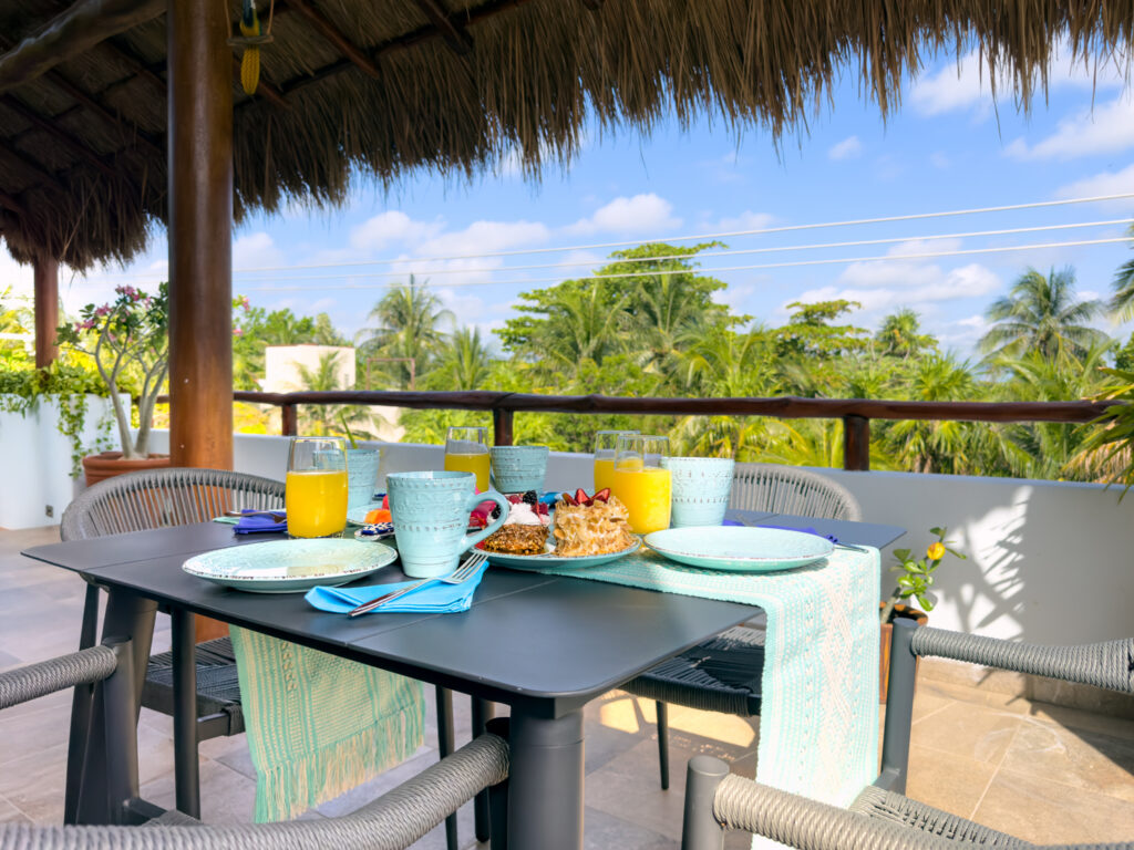 Table set with dishes and drinks beneath a thatched terrace roof overlooking green treetops.