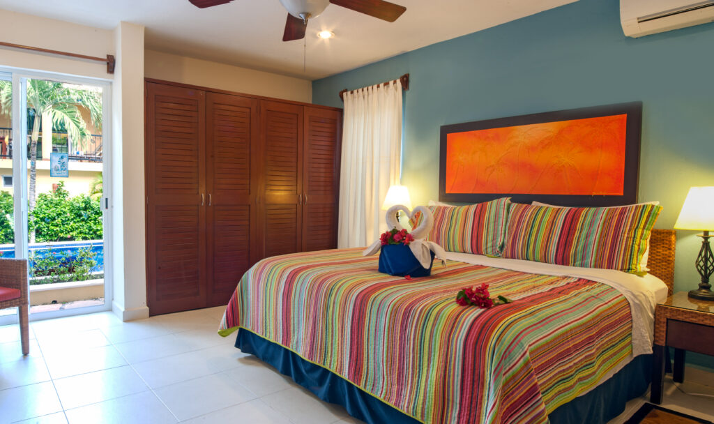 Striped bedspread beneath orange artwork on a blue wall beside an open balcony door.