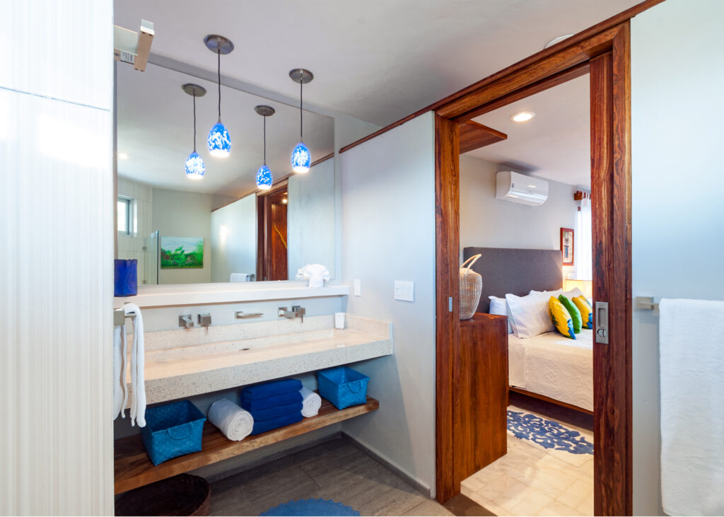 Long vanity with blue pendant lights and open towel storage beside a bedroom doorway.