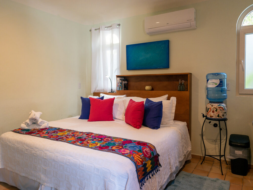 Bed with bright cushions beneath blue artwork, with a water dispenser beside an arched window.