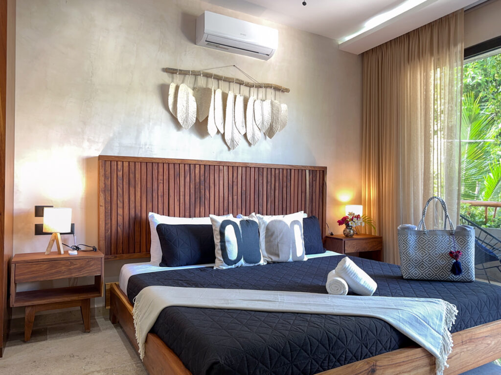 Bed with dark bedding and rolled towels beneath a wooden headboard and decorative wall hanging.