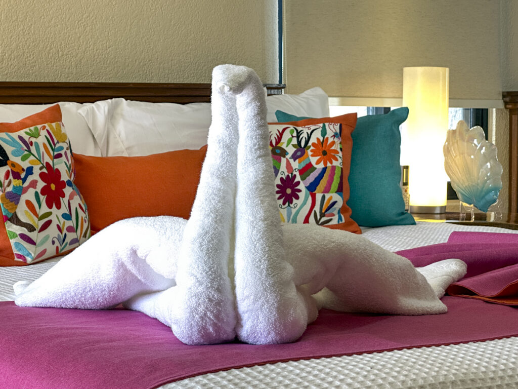 White towel sculpture arranged on a pink bed throw in front of colorful cushions.
