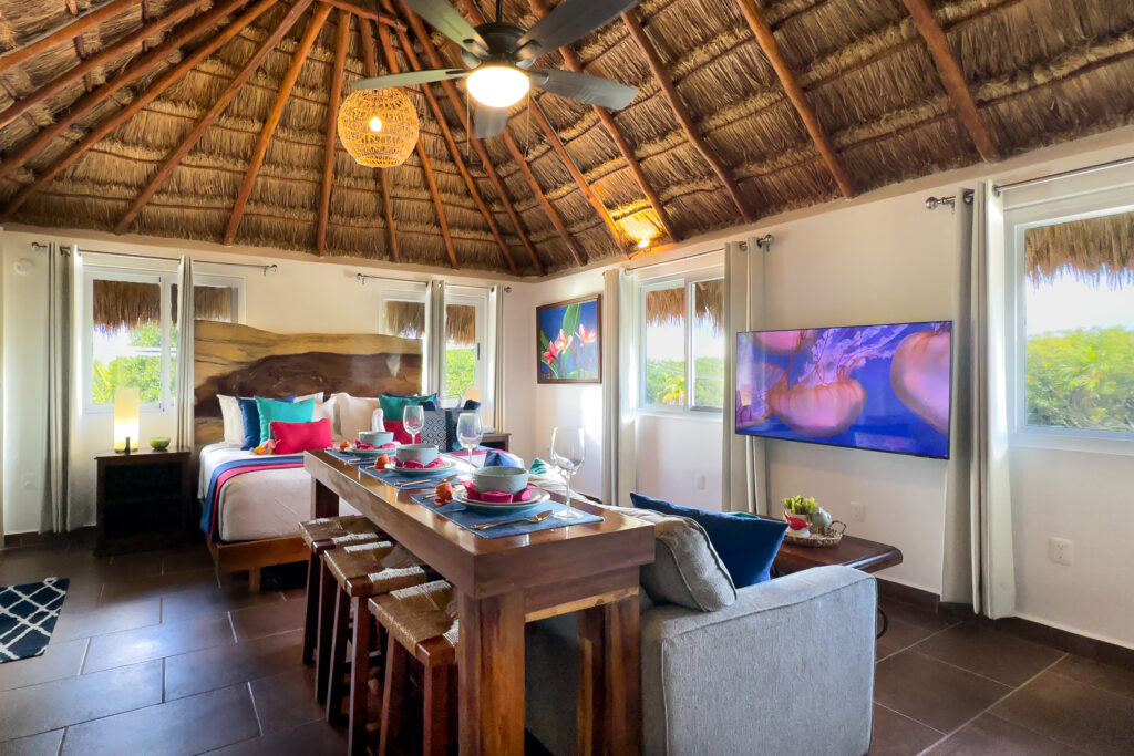 Wooden dining bar separates the sofa and bed beneath a thatched ceiling with a fan.