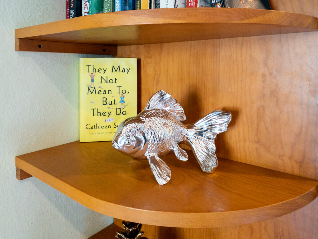 Silver-colored fish ornament on a curved wooden shelf beside books.
