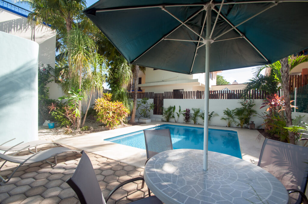 Round outdoor table beneath a large umbrella beside the swimming pool and lounge chairs.