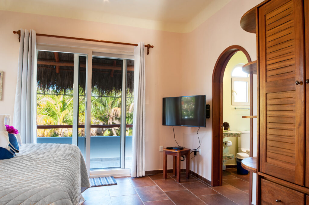 Bed facing a television and arched bathroom doorway beside balcony doors and wooden storage.