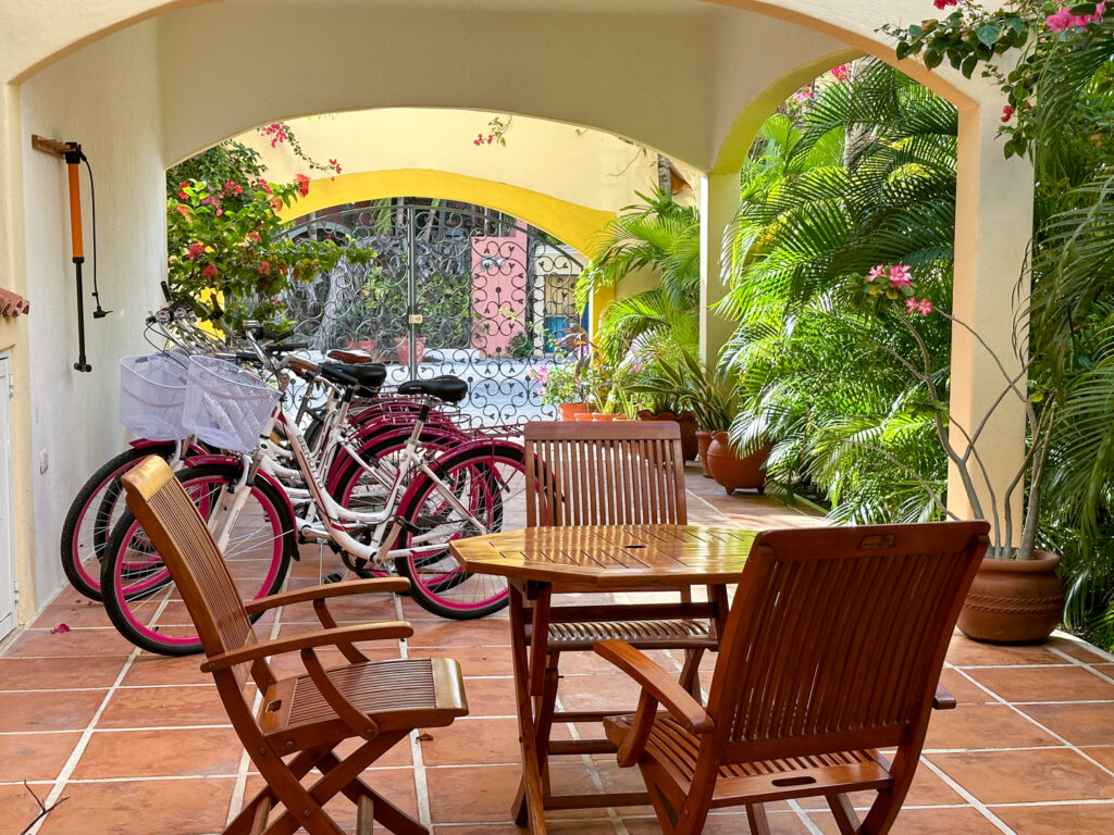 Wooden dining table and chairs beside bicycles beneath yellow patio arches and tropical plants.