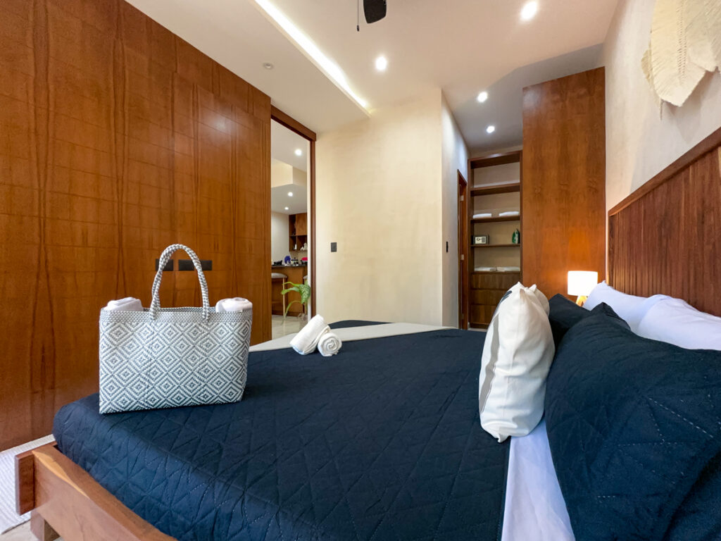 Bed with dark pillows and a woven bag beside a long wooden storage wall leading toward the hallway.