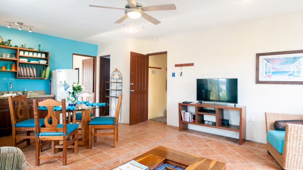 Wooden dining table with blue-cushioned chairs beside a television and wicker seating.